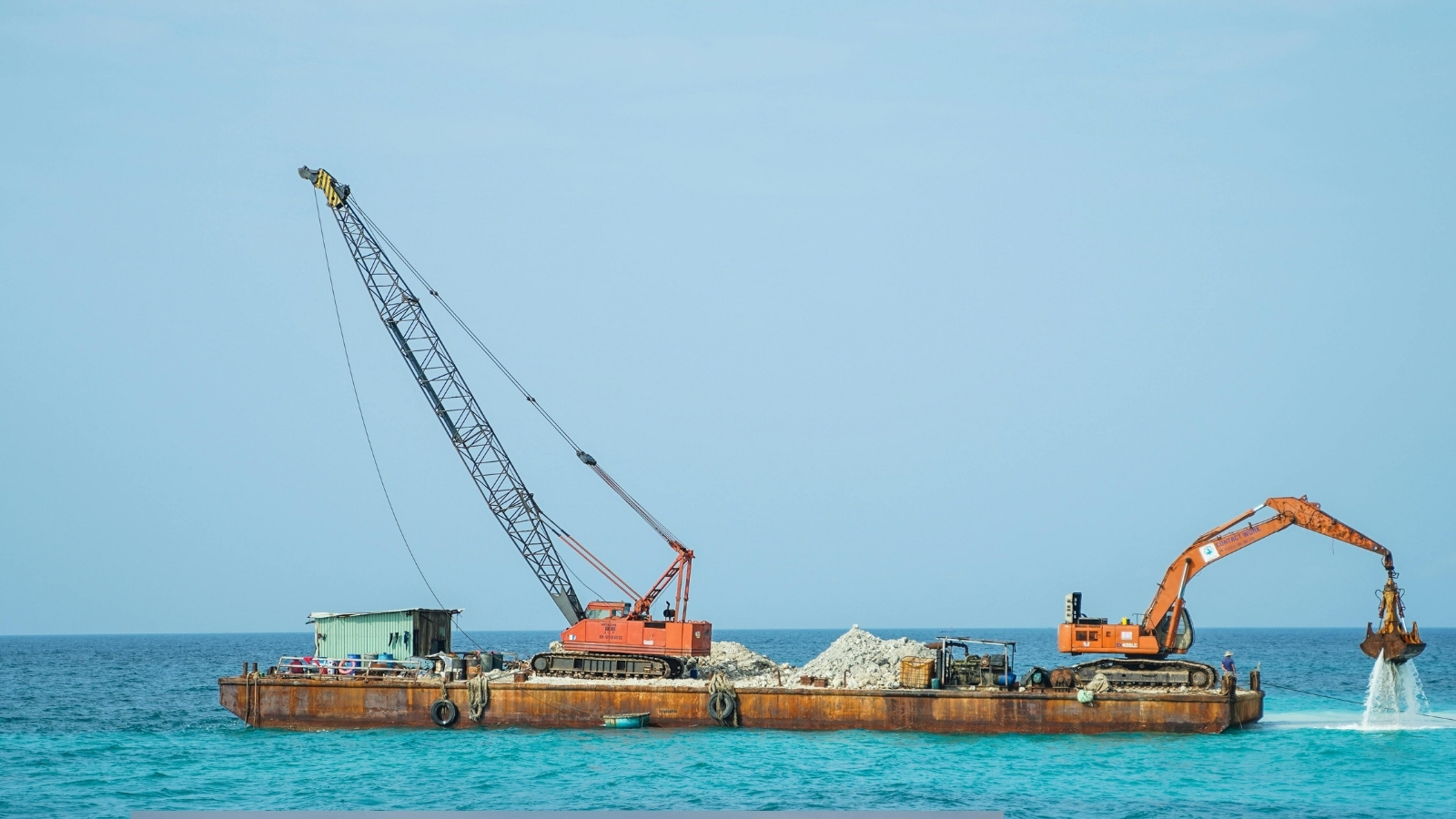 A crane and excavator working from a barge, with the bucket dripping water back into the sea.