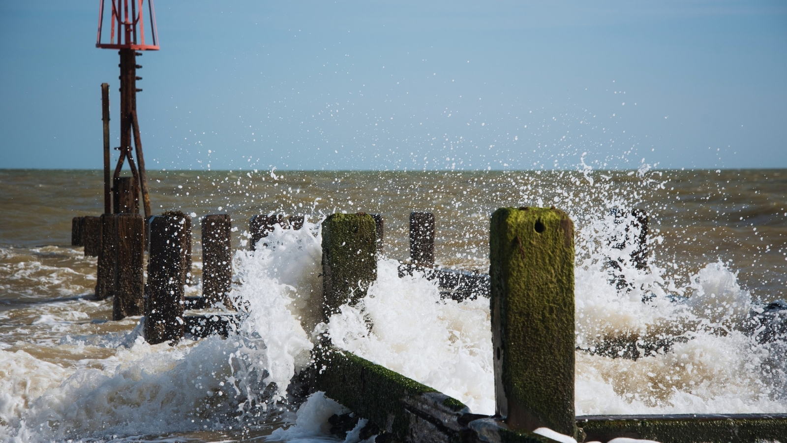 Waves breaking over timber groyne piles, with a red navigation beacon beyond.