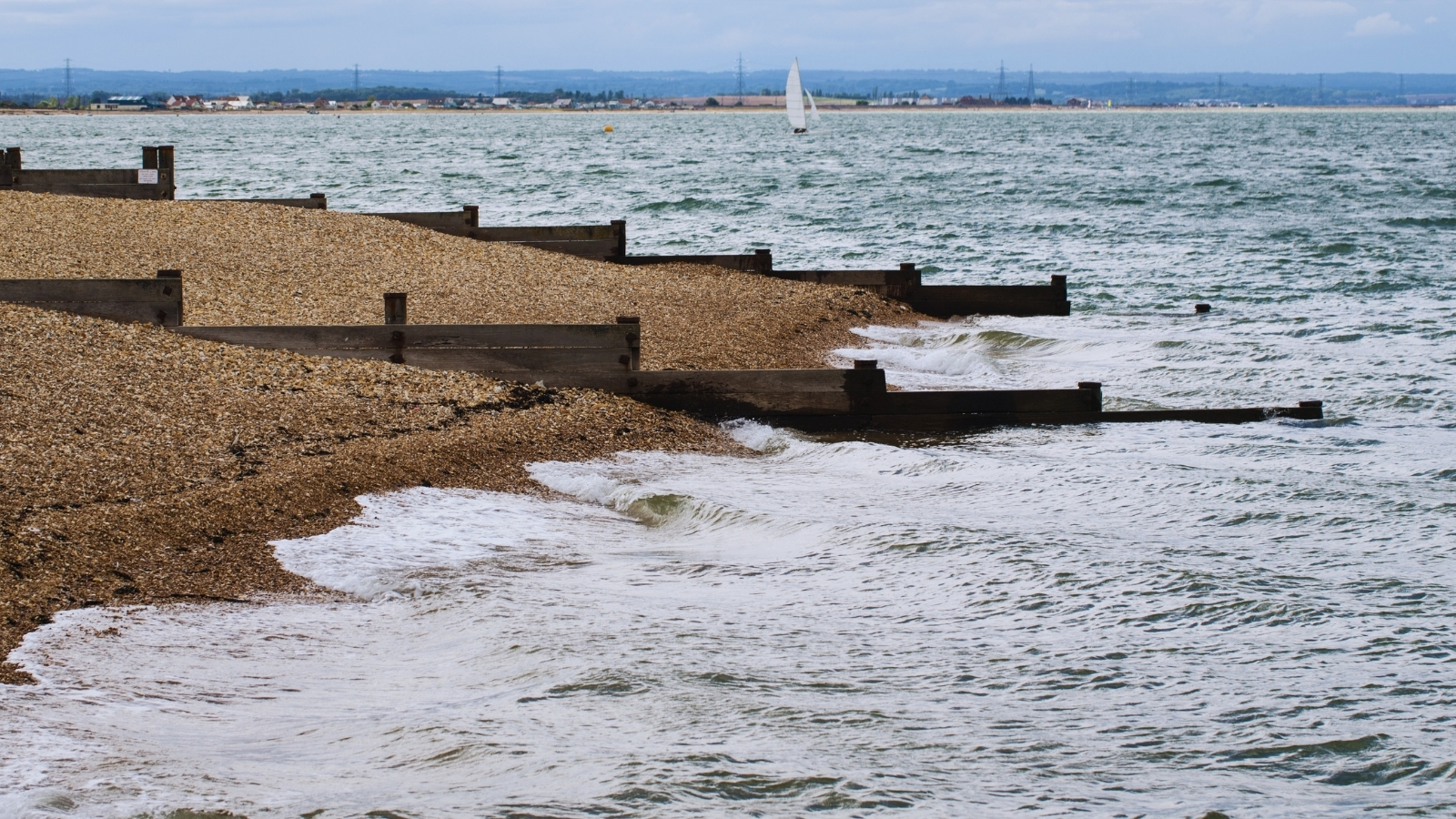 Timber groynes holding a shingle beach, with a yacht sailing offshore.
