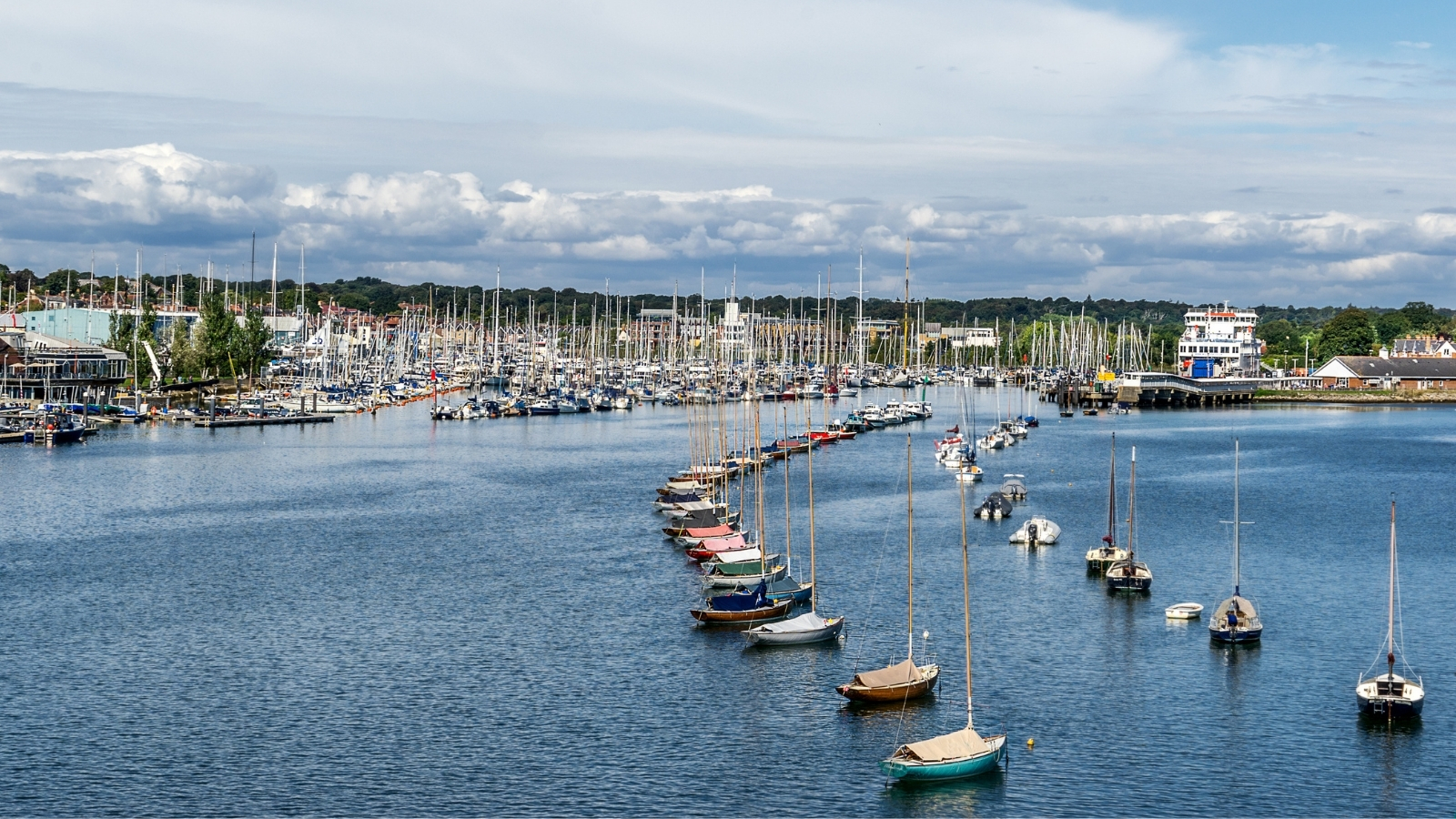 Yachts moored along a curved pontoon in a busy marina, with a ferry at the quay.