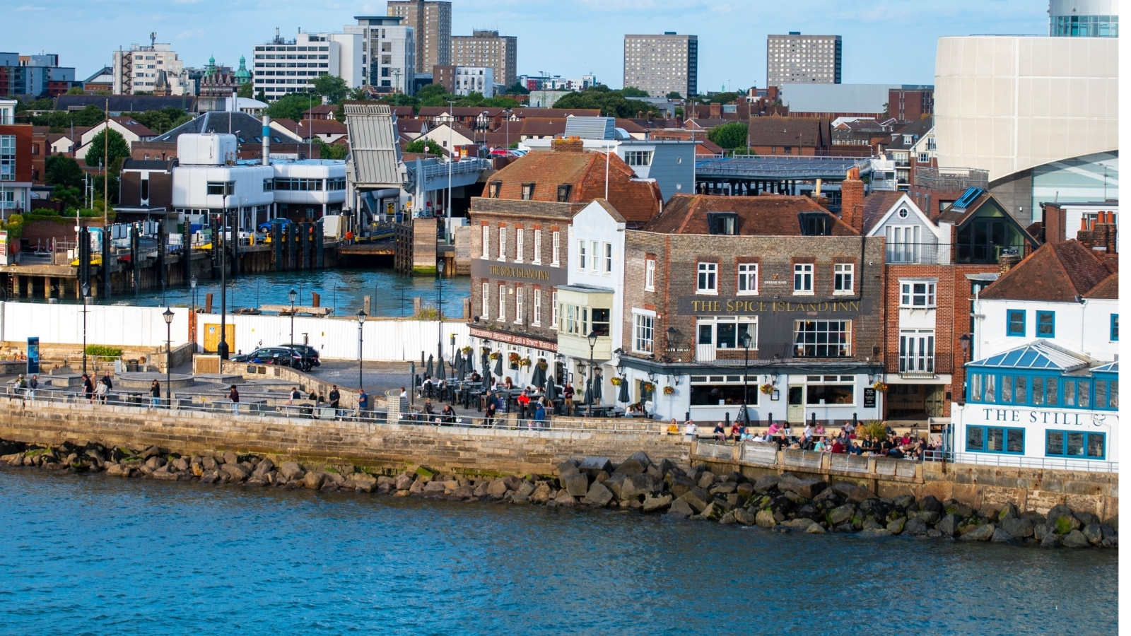 A stone quay wall and rock armour along a waterfront, with the Spice Island Inn behind.